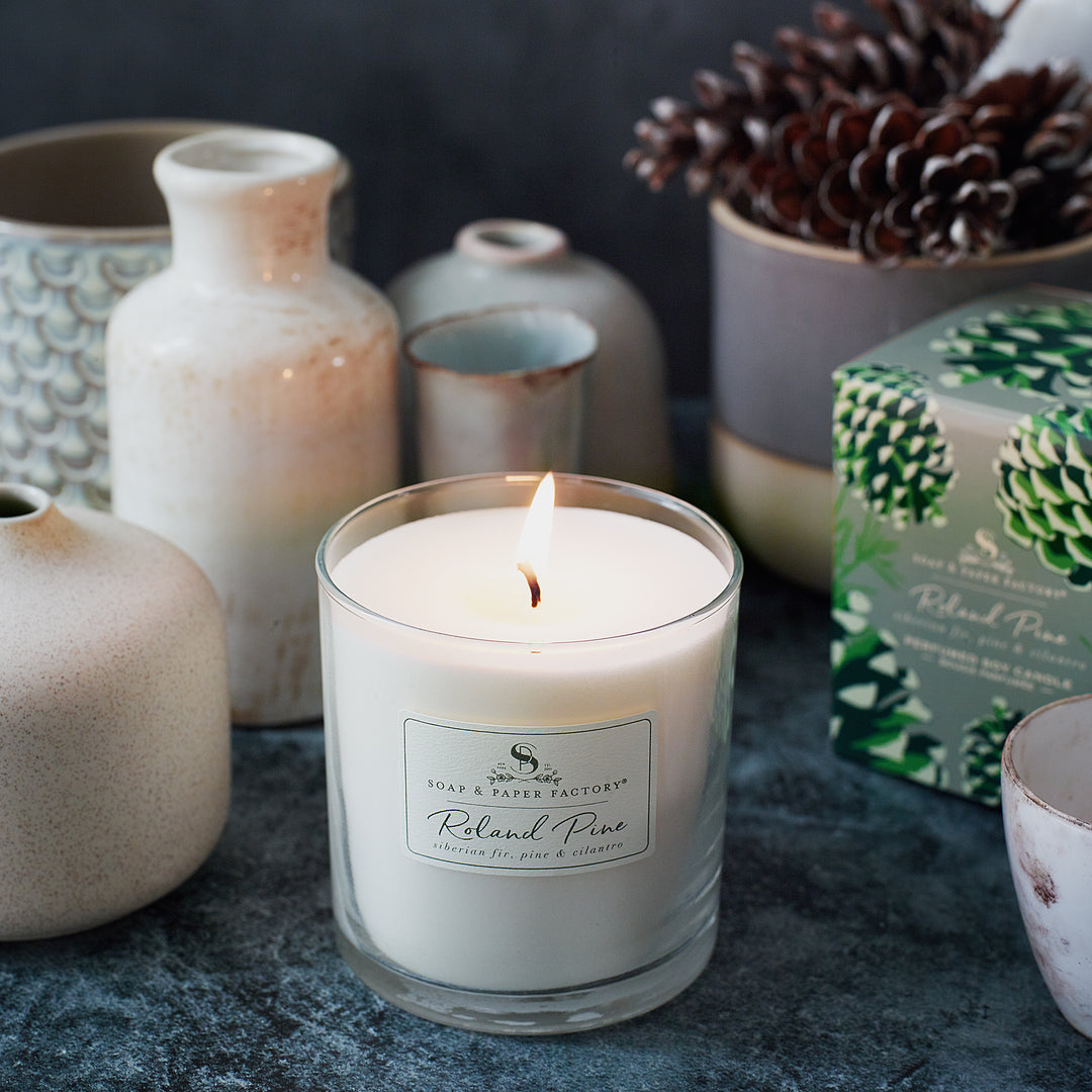 Roland Pine Candle with a label surrounded by decorative vases and pinecones on a dark surface