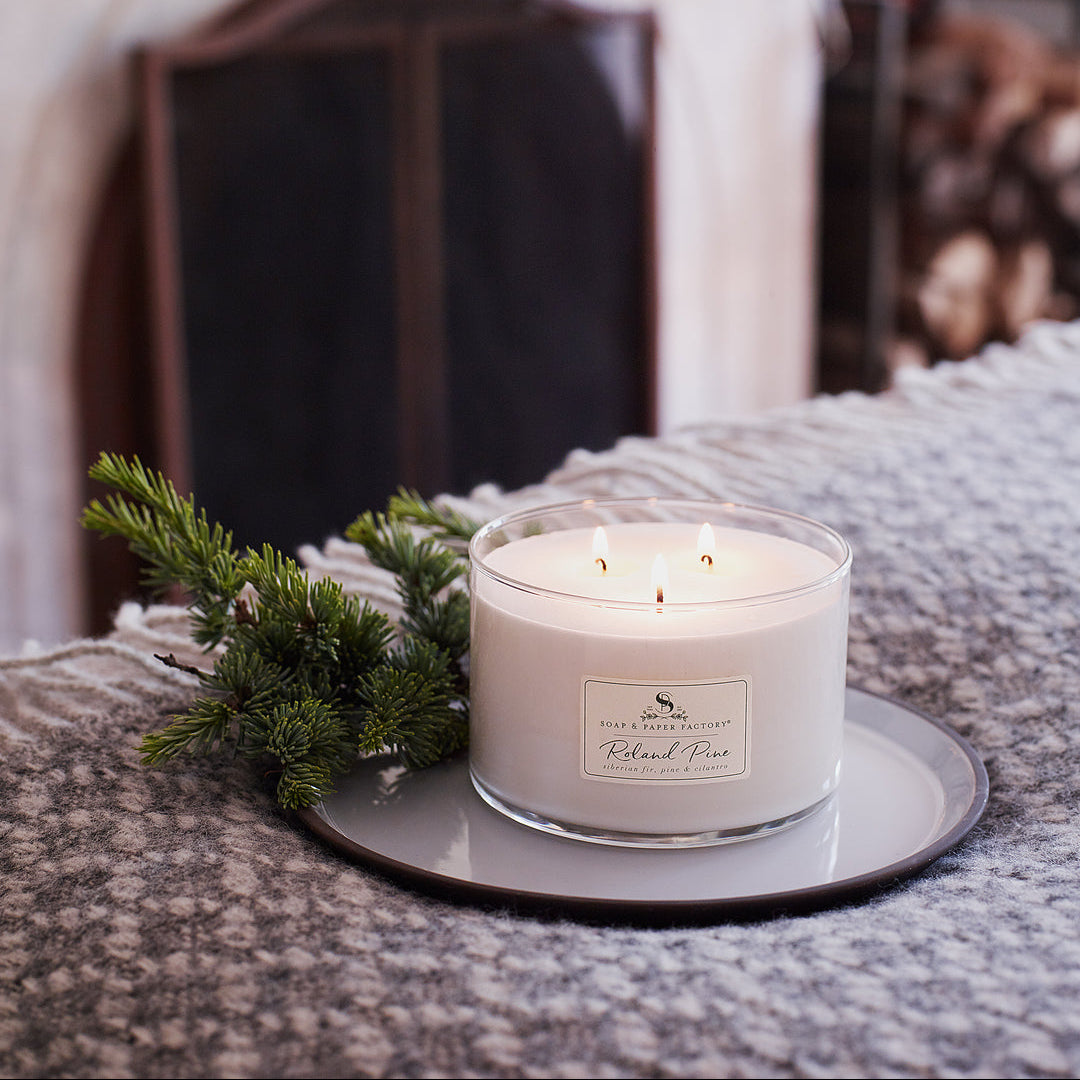 Roland Pine Candle with a label on a small round tray, surrounded by greenery, with a blurred background of lights and a fireplace.