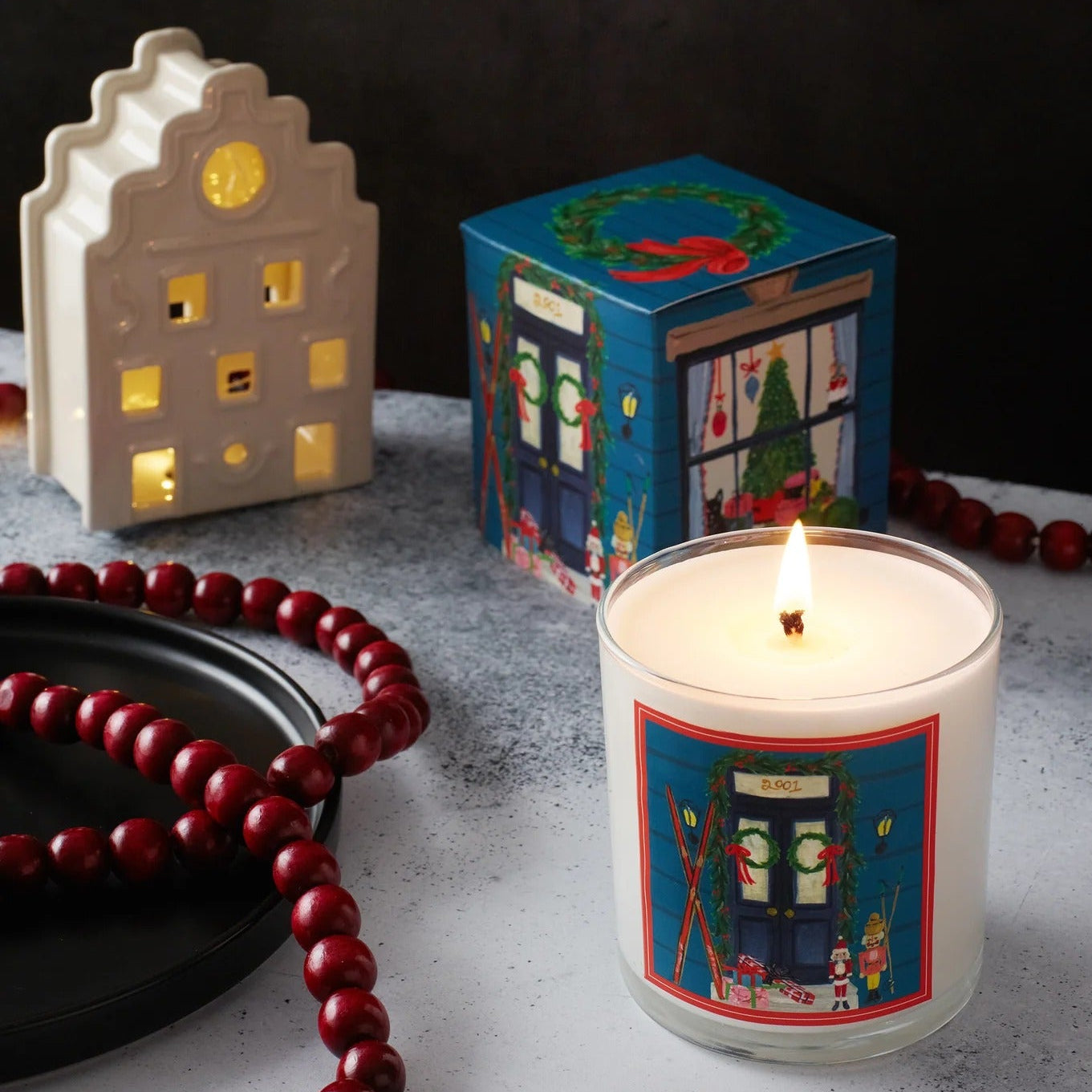 Roland Pine Candle with decorative label, small building lantern, and red beads on a dark background
