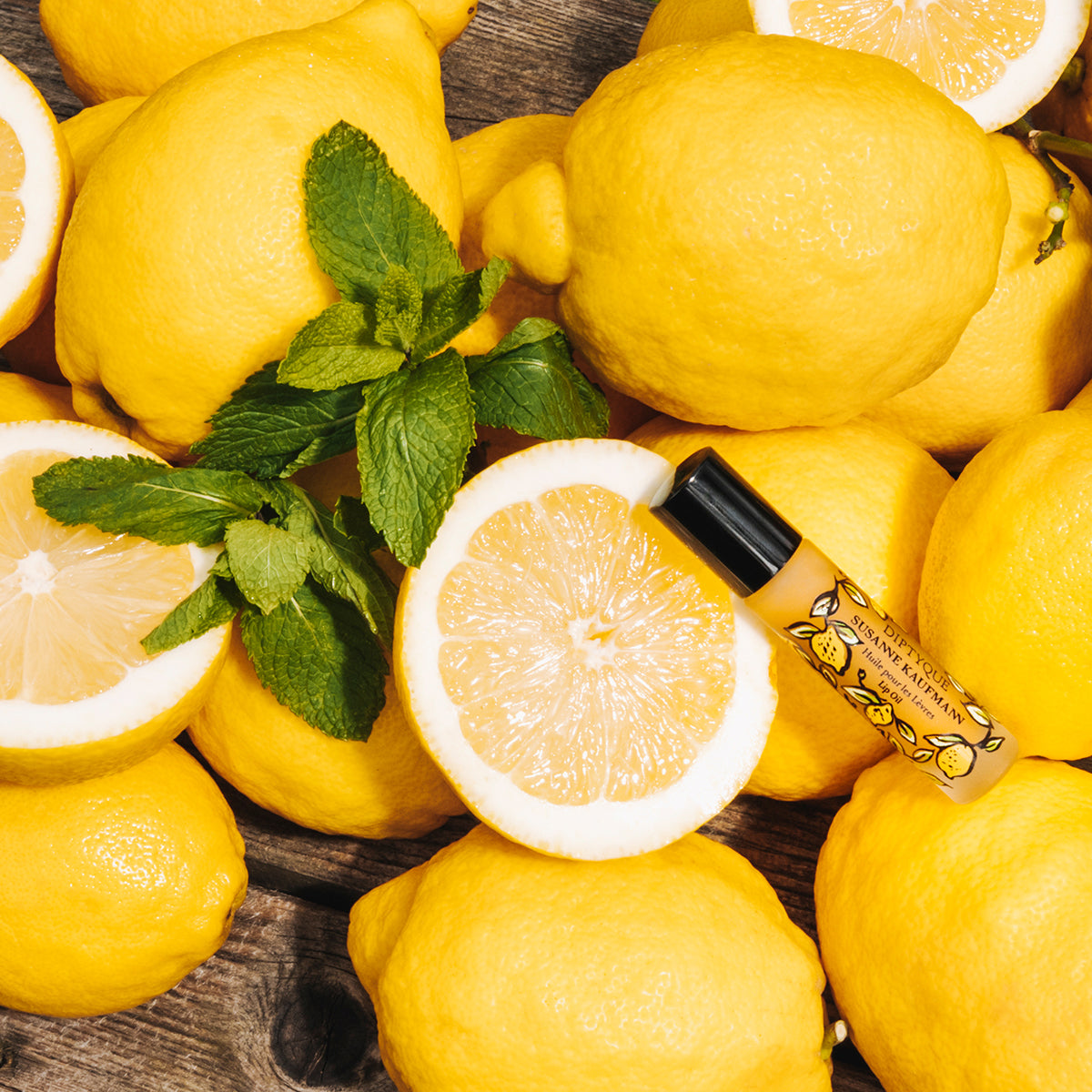 Lip oil surrounded by lemons and mint leaves on a wooden surface