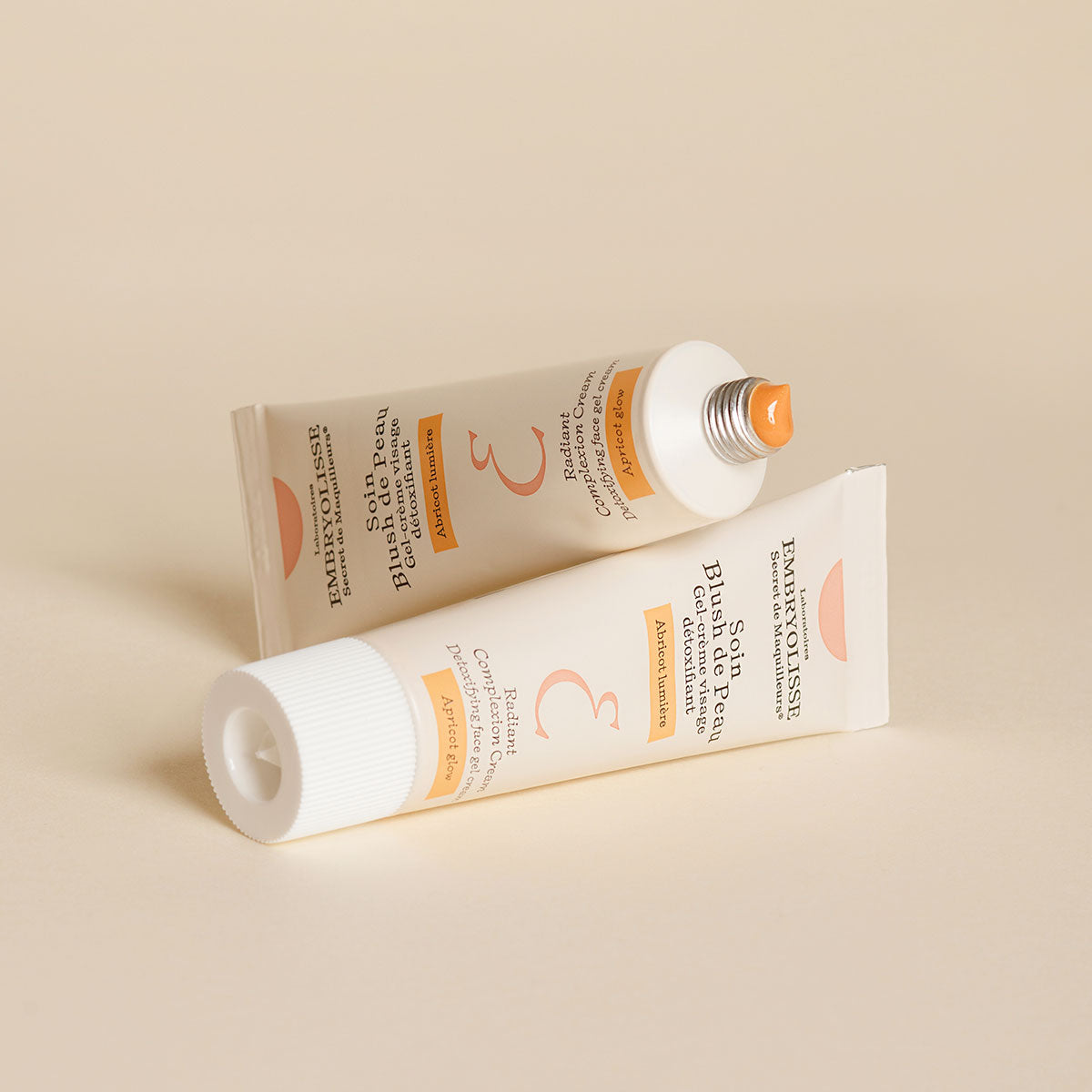 Two bottles of Embryolisse Radiant Complexion Cream in Apricot Glow on tan background.