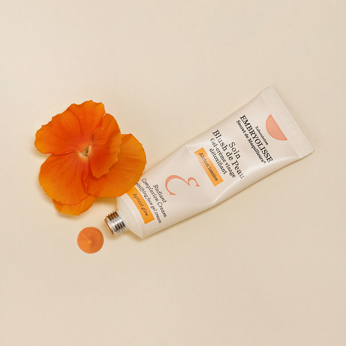 Embryolisse Radiant Complexion Cream in Apricot Glow next to a swatch of product and an orange flower. 