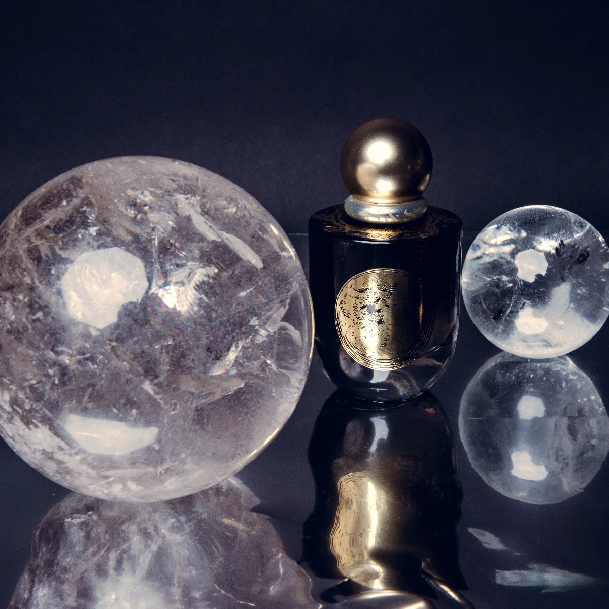 Three decorative spheres on a glossy black reflective surface: a large translucent clear stone ball on the left, a small dark bottle with a metallic gold cap and gold label in the center, and a smaller clear stone ball on the right. The spheres are reflected below.