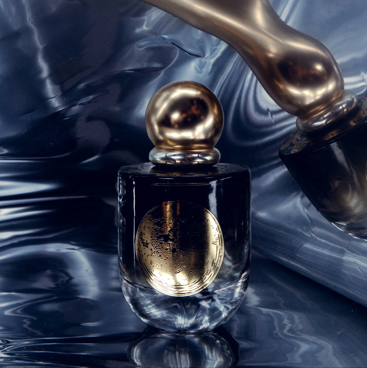 A luxury perfume bottle with a dark glass body, a metallic gold spherical cap, and a gold oval emblem on the front, set against a reflective dark blue and black liquid background.