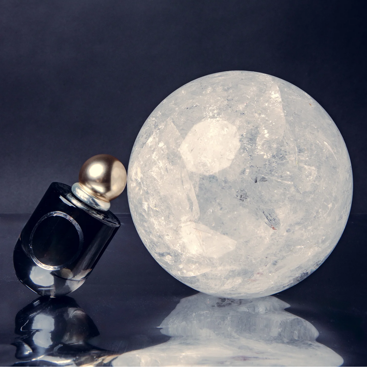 A clear faceted crystal sphere sits beside a dark glass perfume bottle with a gold cap on a reflective dark surface against a dark background.