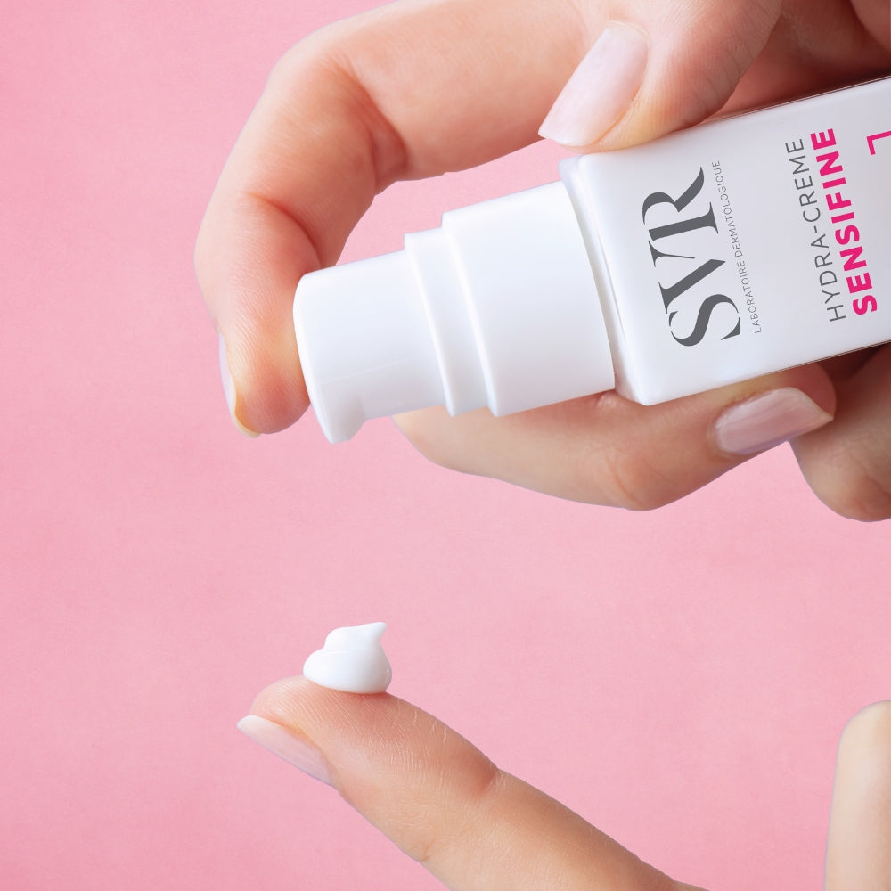 Hand applying SVR Sensifine Hydracream skincare product on a pink background