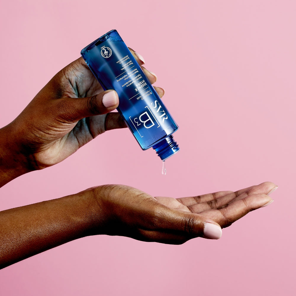 Hand holding a SVR B3 Hydra Essence  bottle of skincare product against a pink background