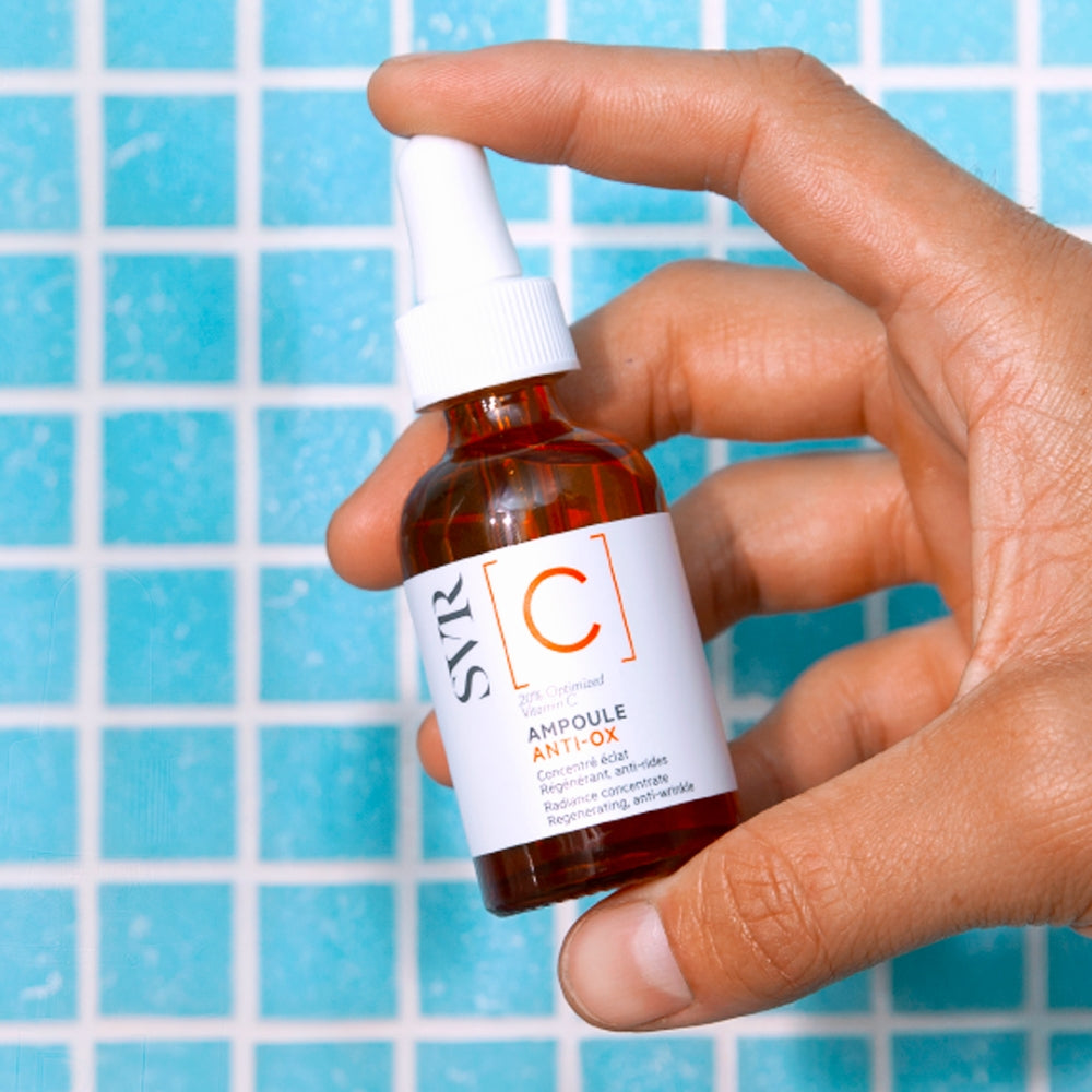 Hand holding a bottle of SVR C Radiance Concentrate serum against a blue tiled background