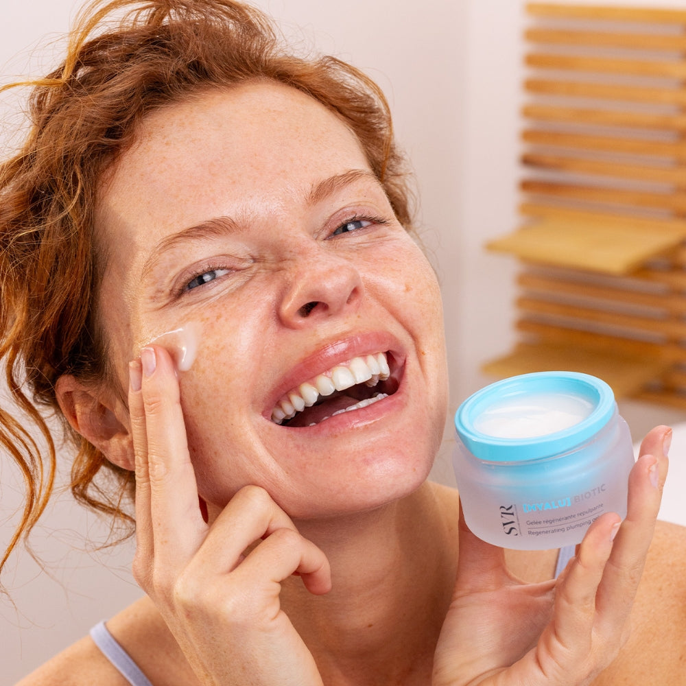 Woman applying cream to her face with a jar of SVR Hyalu gel cream in her hand, set against a neutral background.