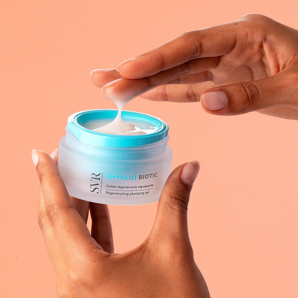 Hand applying cream from a jar labeled SVR Hyalu Biotic Plumping gel against a peach background