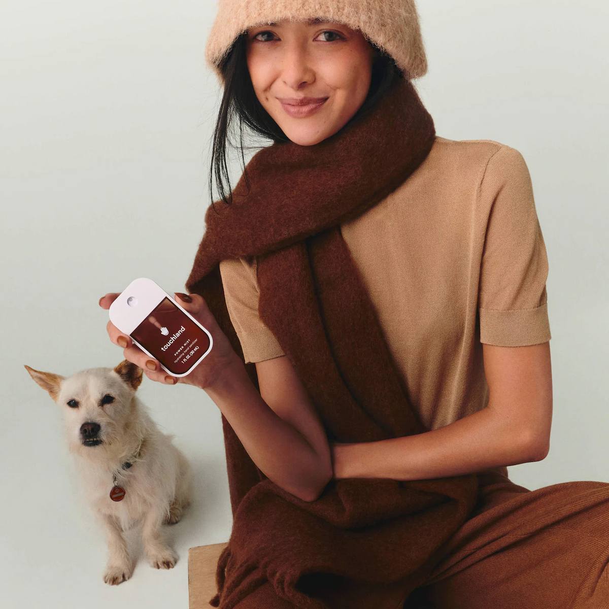 A woman wrapped in a brown knit scarf and tan top, wearing a beige beanie, holds a small white dog and a smartphone showing a brown pet tracking app screen. The dog wears a matching brown collar with a tag.