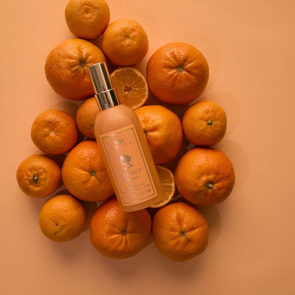 A bottle of TOCCA Sea Salt Riviera - Hair and Body Mist is arranged on top of a circular cluster of fresh oranges against a solid peach background.