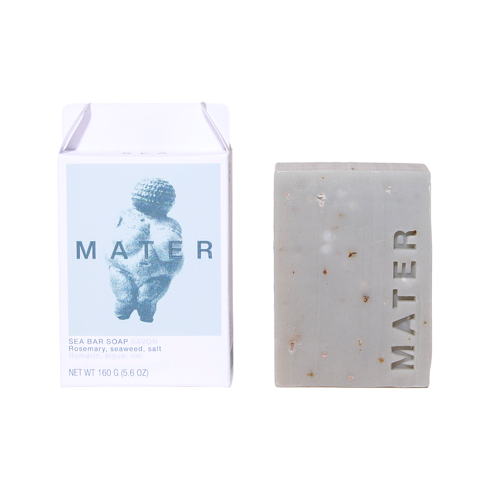 A bar of MATER Sea Bar Soap next to its white packaging box on a clean white background. The soap is a light gray with tiny embedded sea salt crystals, and the box features a light blue graphic of a cherub with the brand name MATER.