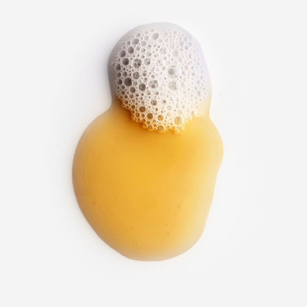A dollop of translucent golden yellow liquid with a frothy white foam top, photographed on a clean white background.