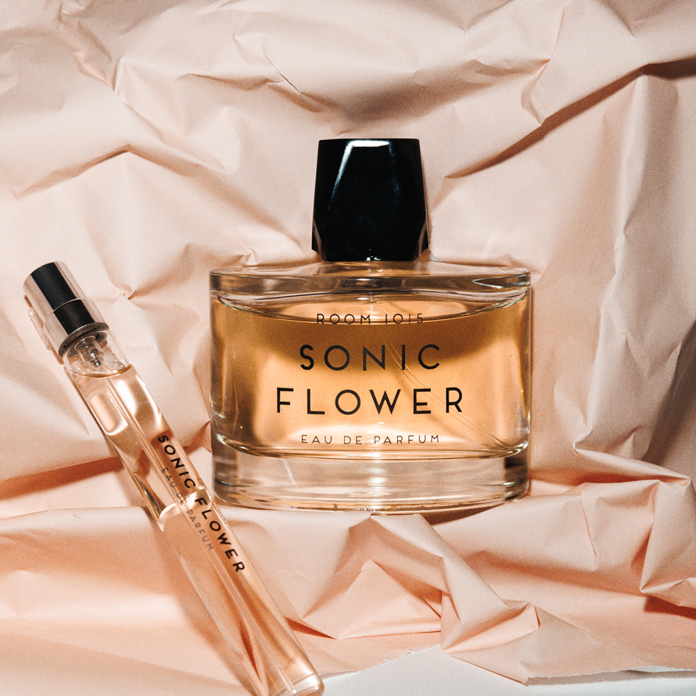 A bottle of Sonic Flower Eau de Parfum with a black cap sits on crumpled blush paper next to a matching clear glass travel spray vial labeled 'ROOM 1015 Sonic Flower'.