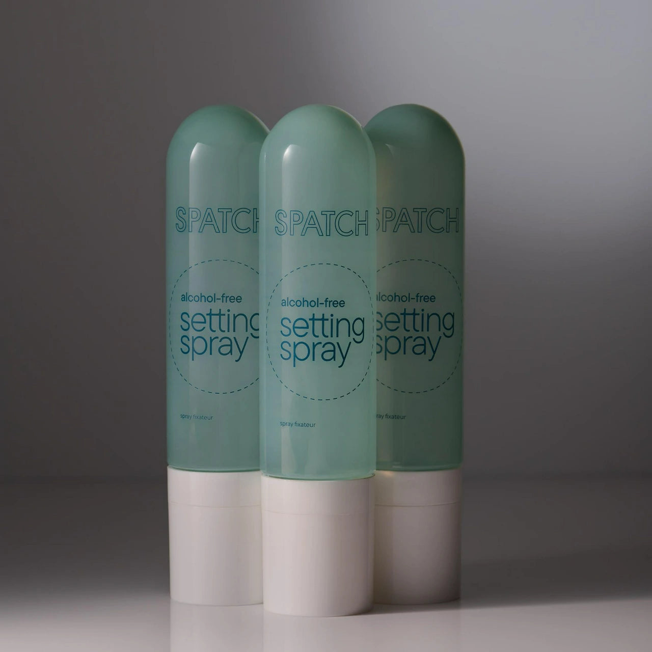 Three bottles of SPATCH alcohol-free setting spray arranged together on a reflective surface against a dark gray gradient background.