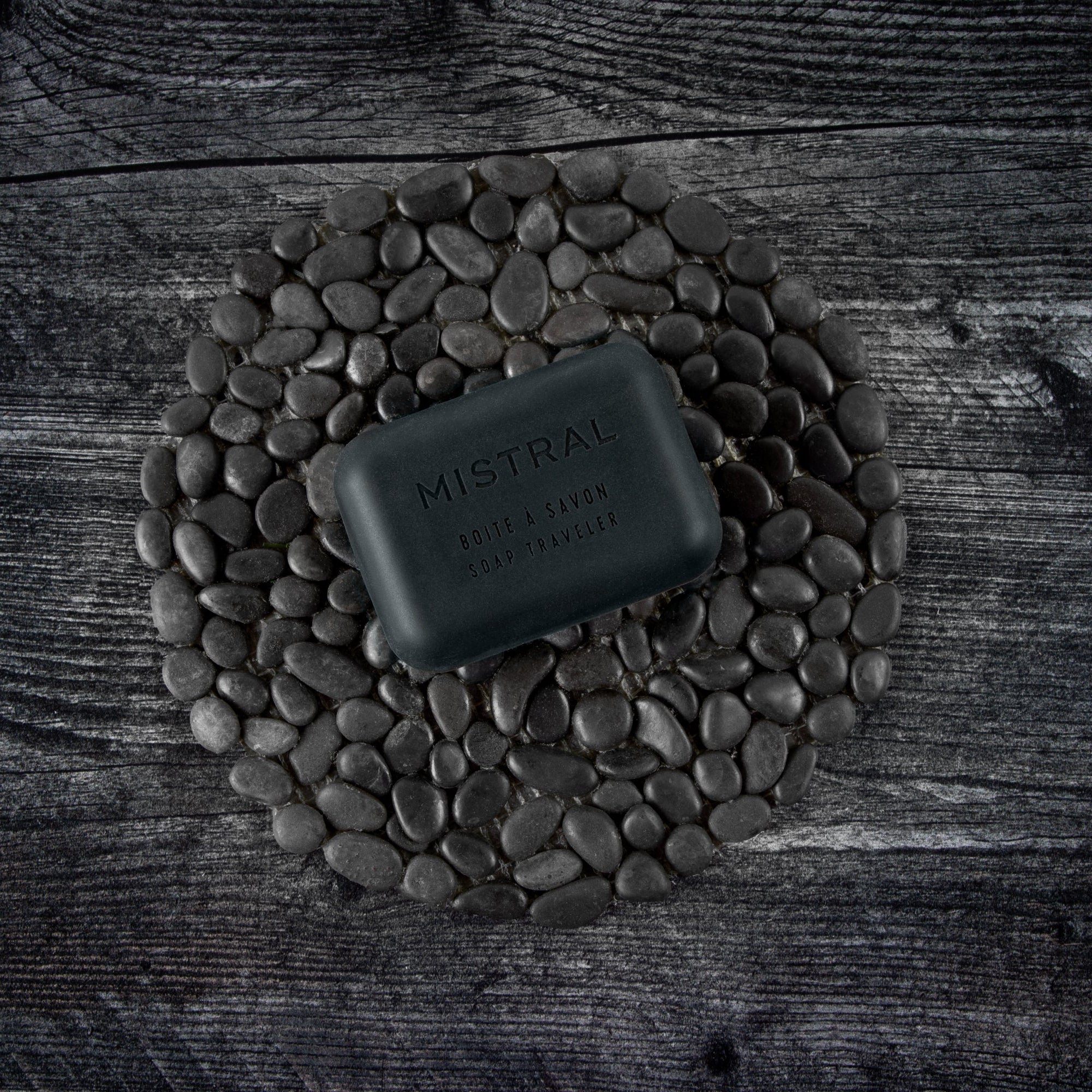 Black soap bar on a circular arrangement of stones with 'MISTRAL' branding on a wooden surface.