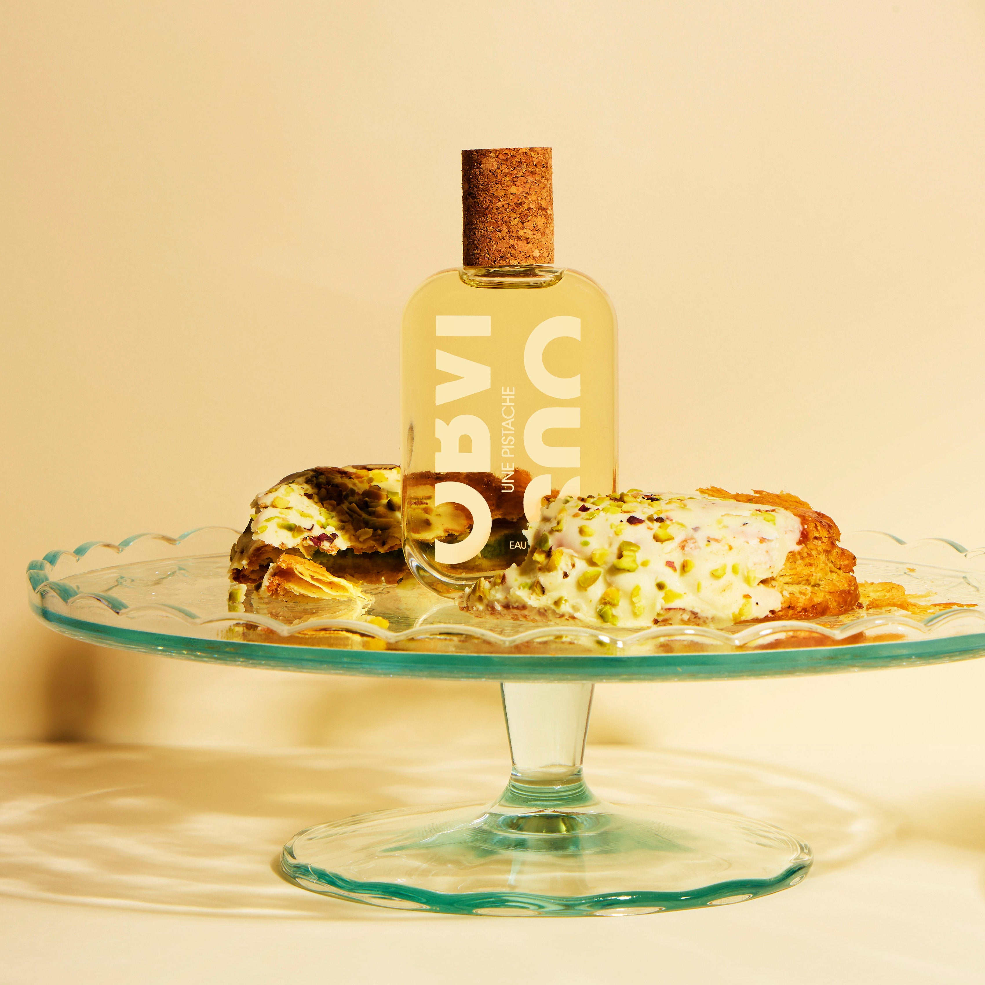 Small bottle with cork and a cake on a glass stand against a beige background