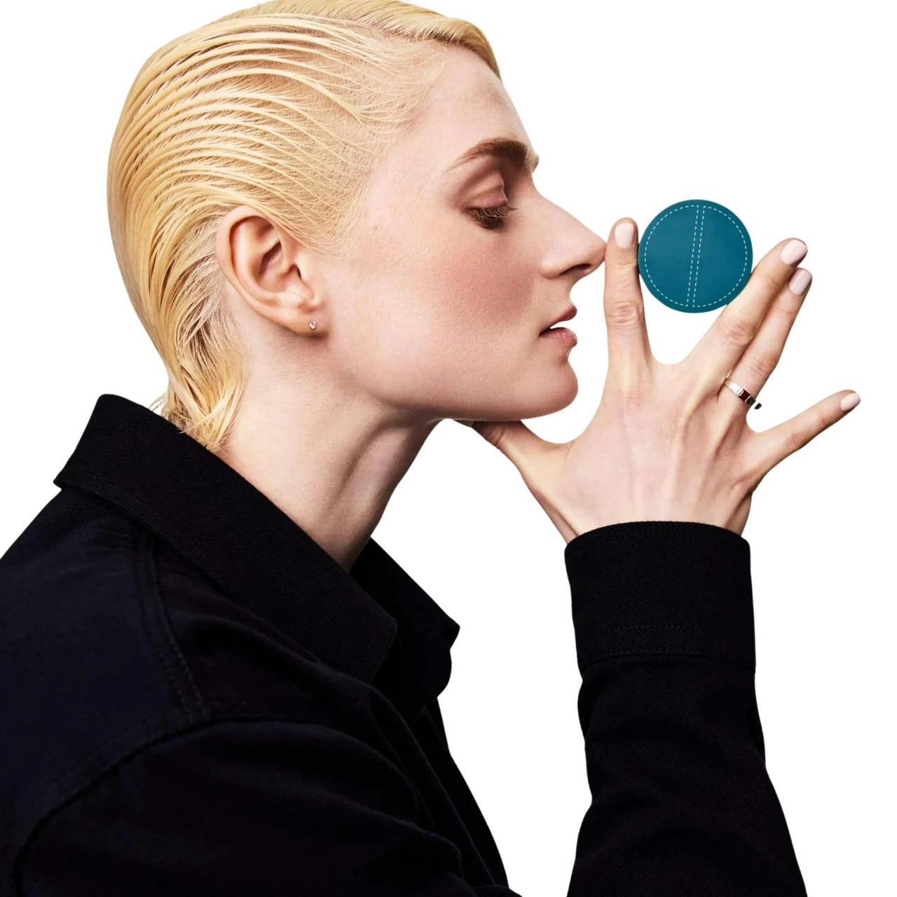 A blonde person in a black collared jacket holds a small round teal ring against their nose with their fingers, shown against a plain white background.