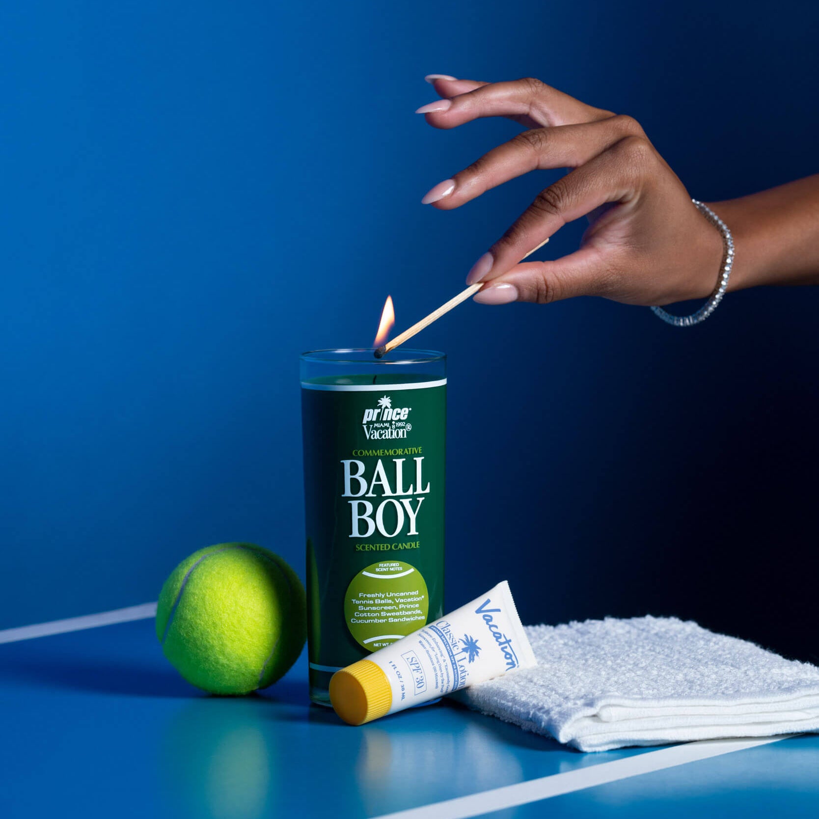 Hand lighting a candle labeled 'Ball Boy' on a tennis court with a tennis ball and towel nearby.