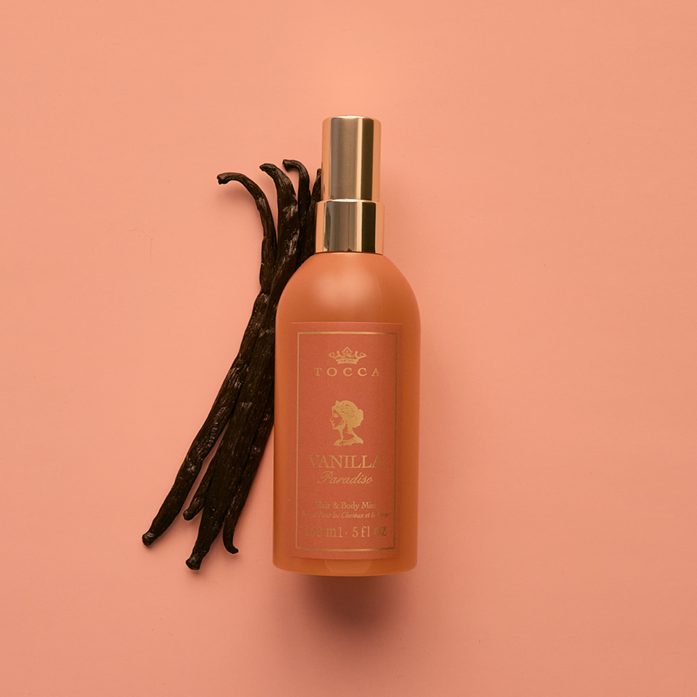 A bottle of TOCCA Vanilla Paradiso - Hair and Body Mist in a frosted peach color with a gold spray nozzle, styled next to vanilla bean pods on a solid peach background.