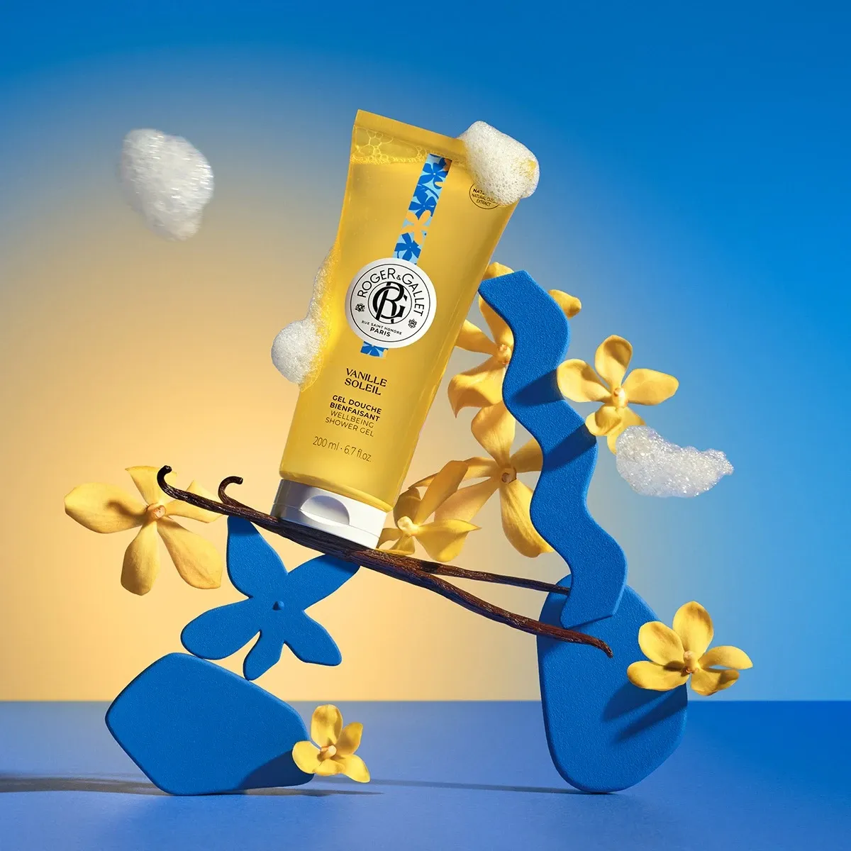 Yellow tube of Roger & Gallet Vanille Soap on a blue and yellow gradient background with flowers and leaves.