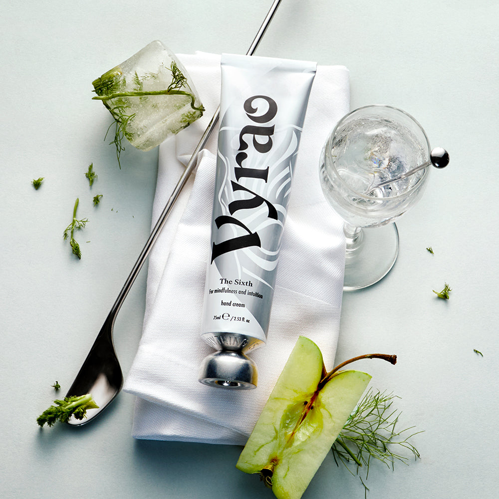 Tube of Vyrao hand cream on a white napkin with a glass of water and apple slices on a light blue background
