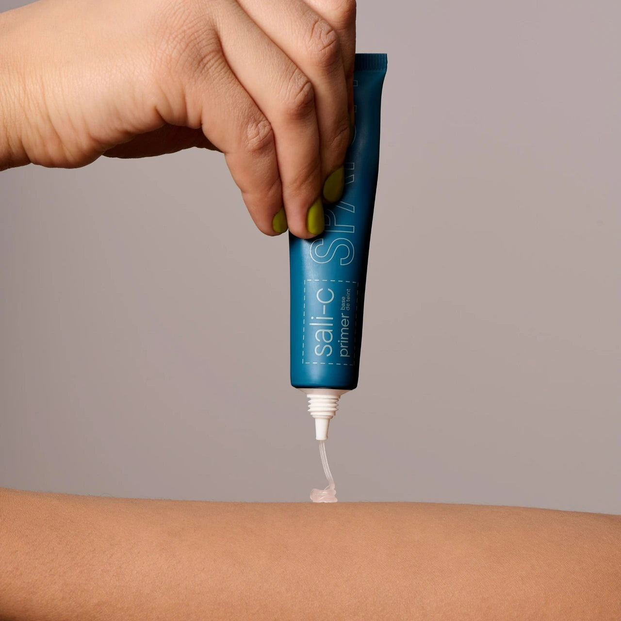 A hand squeezes a blue tube of sali-c hemostatic primer onto a person's forearm, with the white gel being dispensed. The background is a solid, neutral gray.