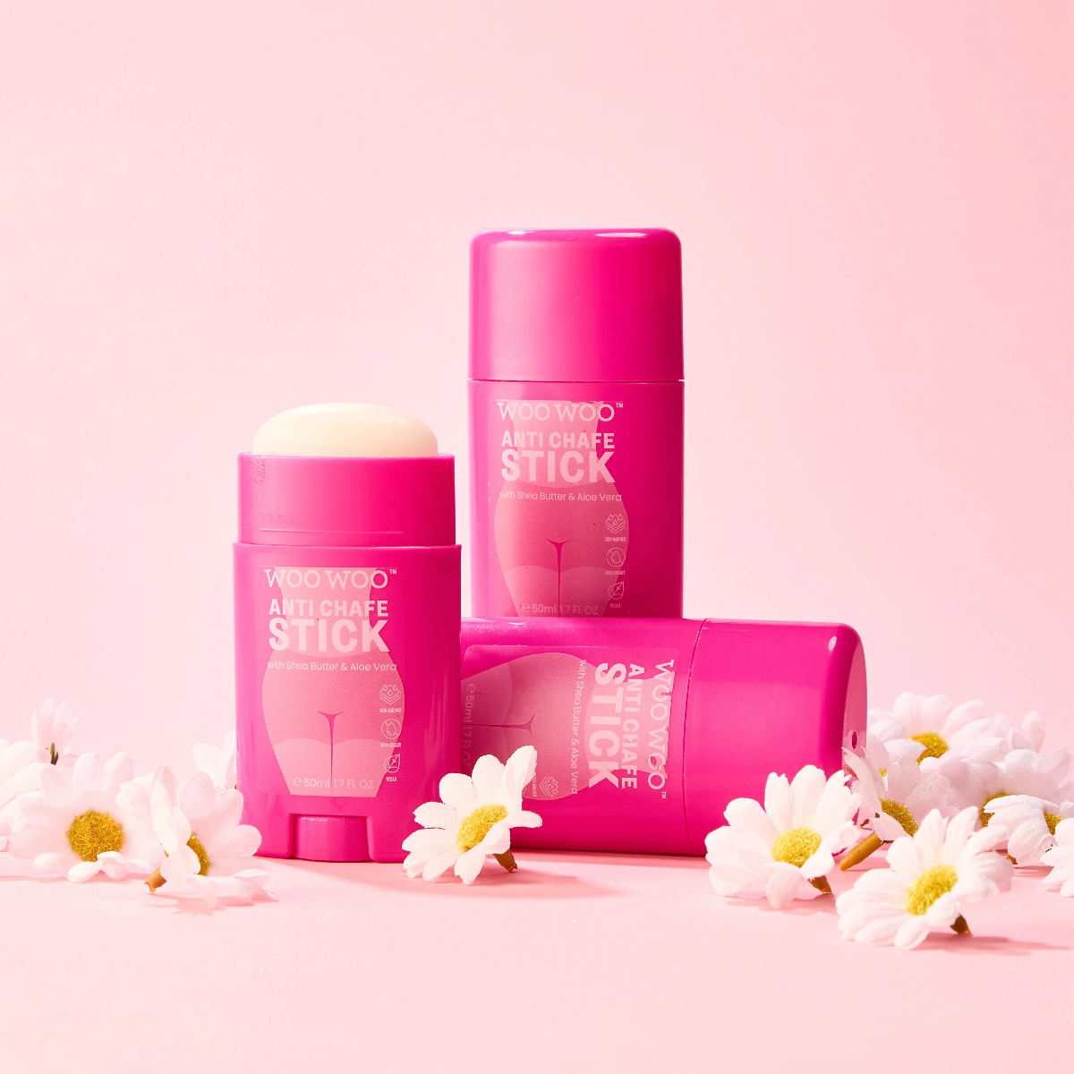 Three pink WOO WOO Anti Chafe Stick deodorant containers arranged on a light pink background with white daisy flowers around them. One stick is upright in front, one is upright behind, and one lies on its side in the middle.