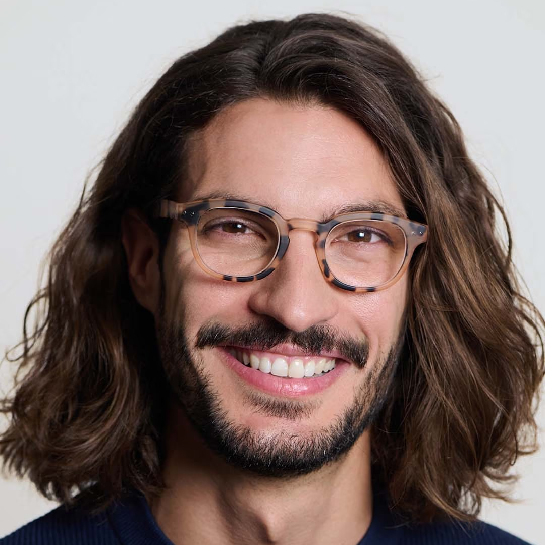 Man with long hair and glasses smiling against a plain background
