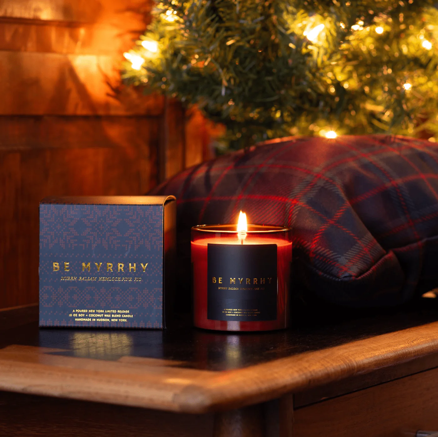 Lit candle with 'Be Myrrh' packaging on a wooden surface with a blurred Christmas tree in the background.