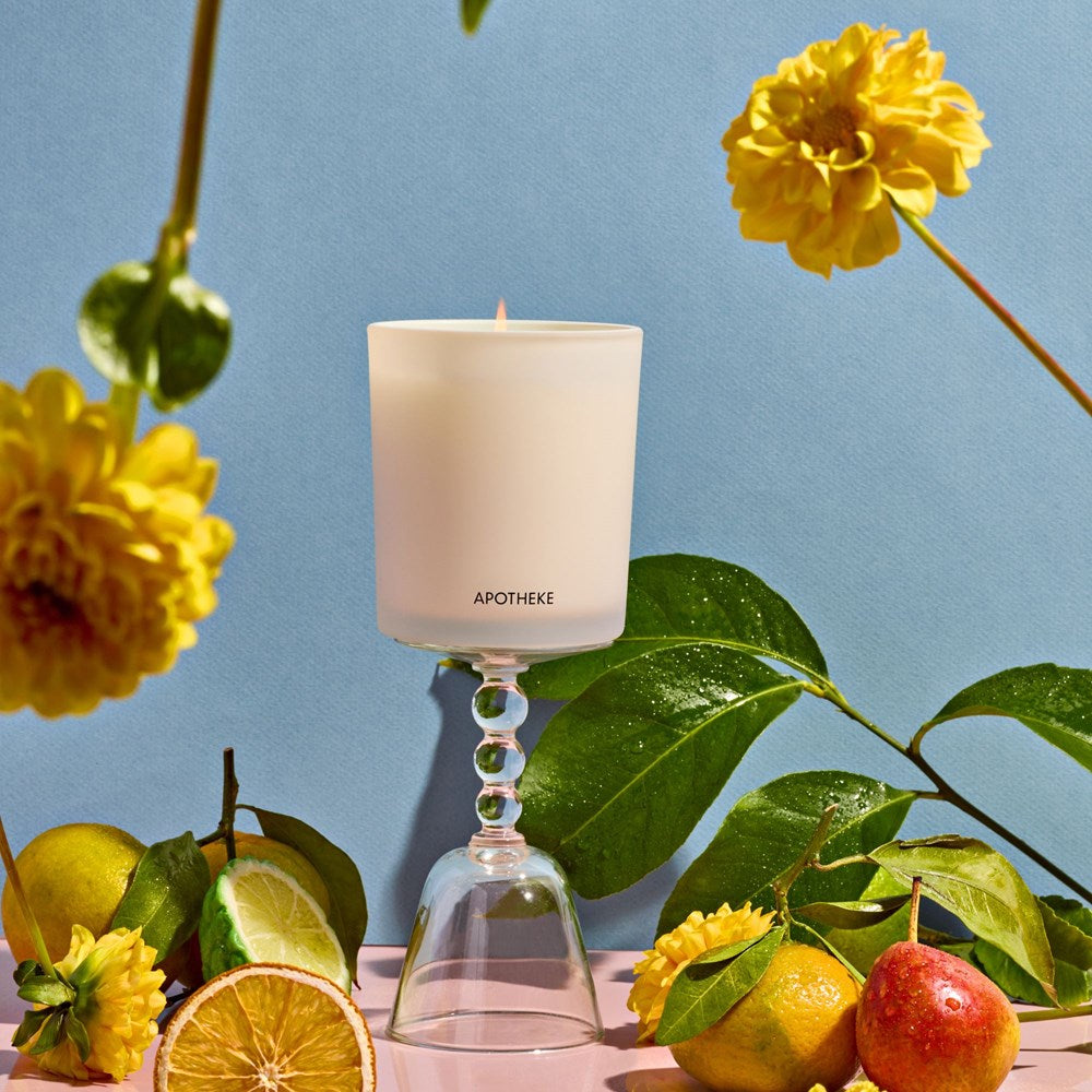 Bergamot Tangerine Candle on a glass pedestal in front of a blue background surrounded by citrus fruits, leaves, and flowers. 