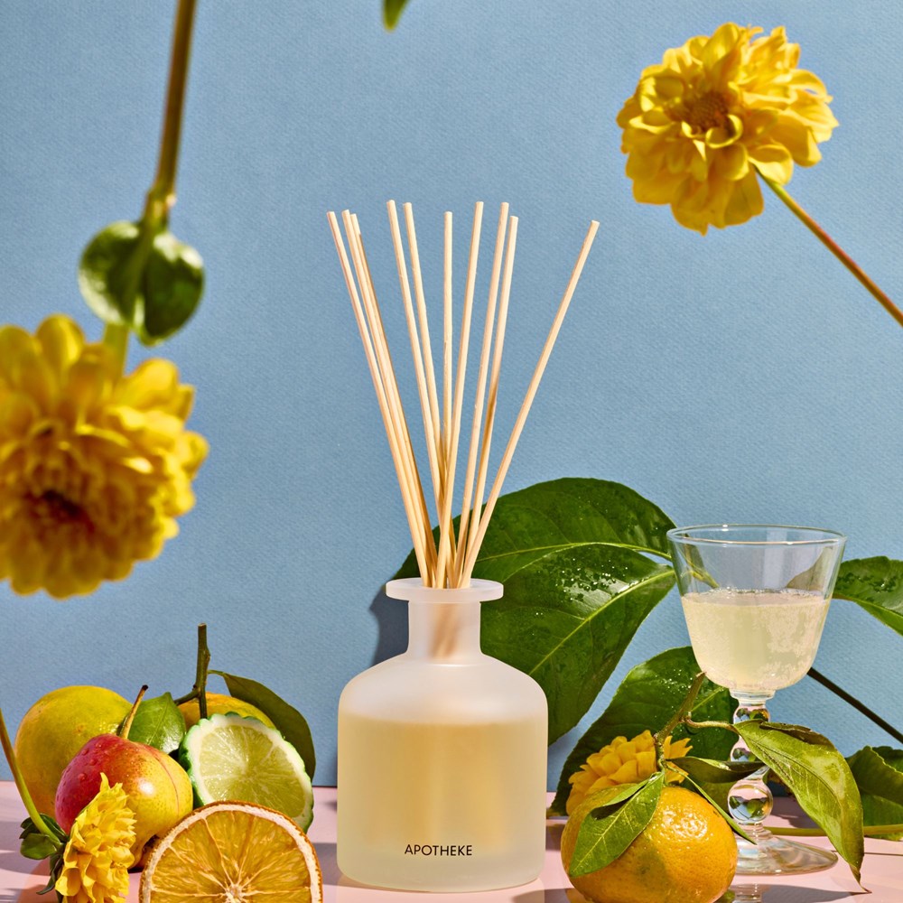 Apotheke Bergamot Tangerine Diffuser in front of a blue background surrounded by citrus fruits and flowers.