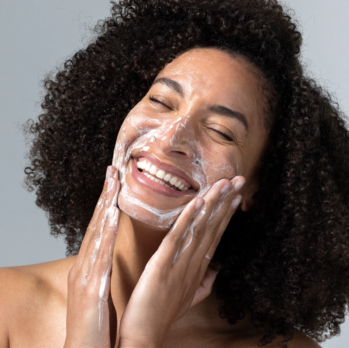 A close-up of a smiling woman with curly dark hair washing her face with white foaming skincare cream against a plain gray background.