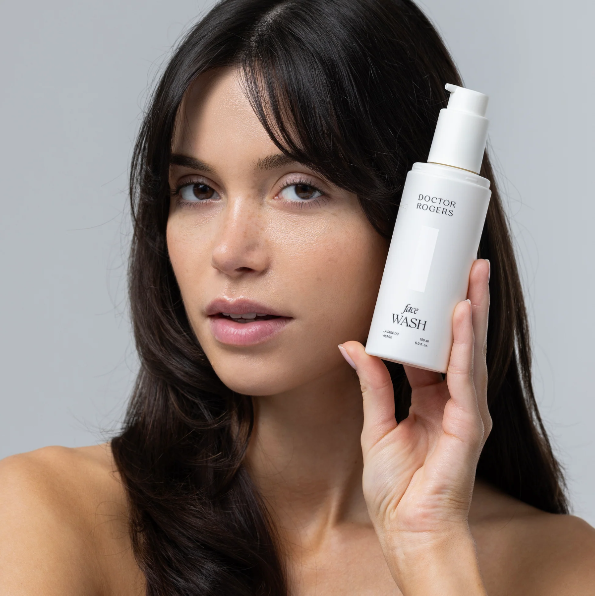 Woman holding a bottle of 'Doctor Rogers' face wash against a gray background
