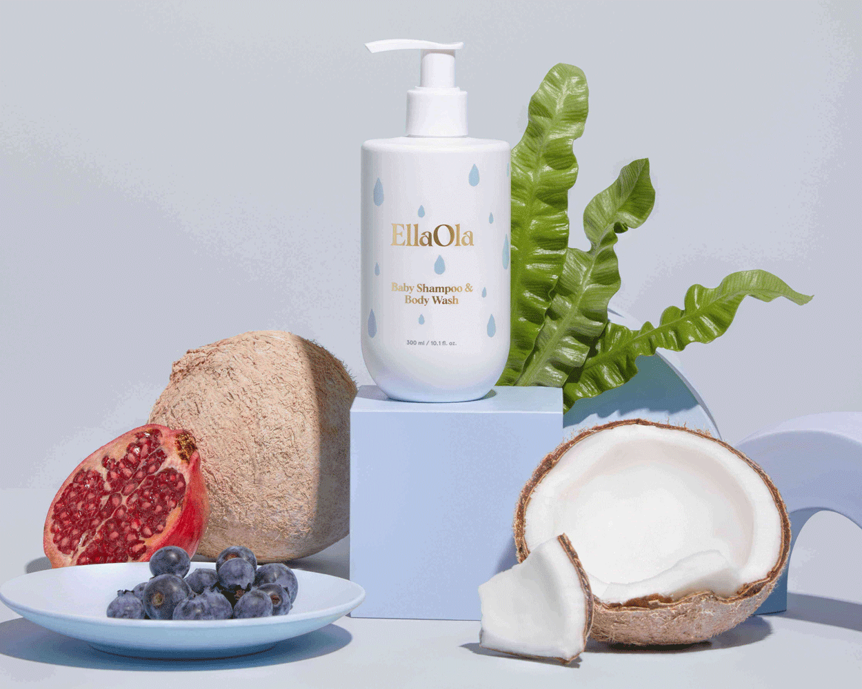 Ella Ola Baby Shampoo and Body Wash on a blue block surrounded by a pomegranate, blueberries, coconut and green leaves in front of a blue background.