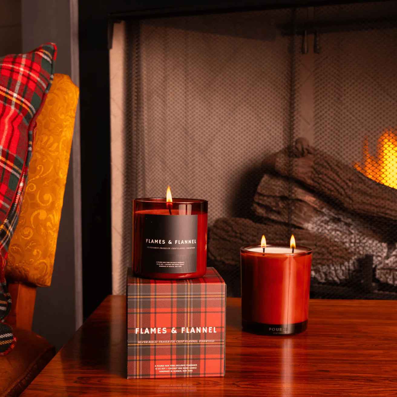 Two red candles with 'Flames & Flannel' branding in front of a fireplace.
