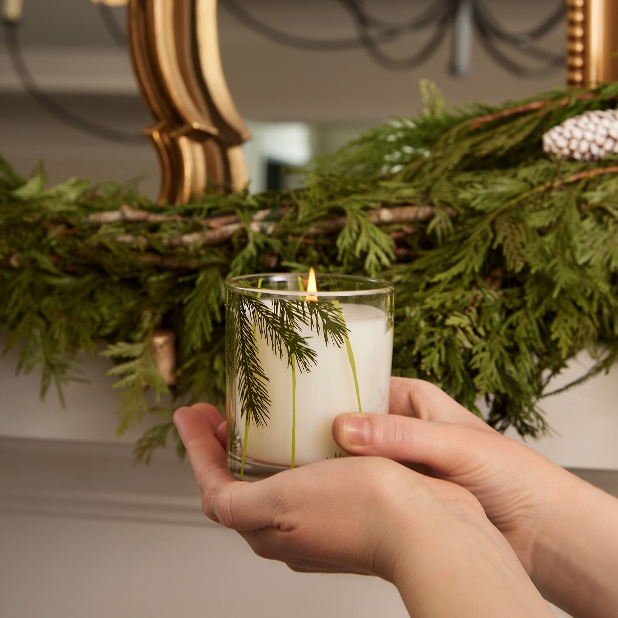 Hand holding a candle with greenery inside, against a decorative background