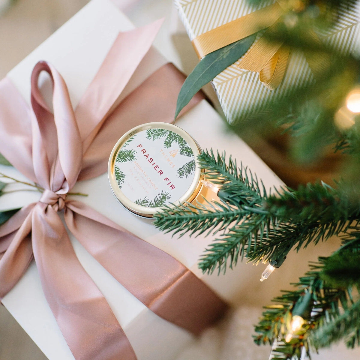 Gift box with pink ribbon and 'Frasier Fir' candle, surrounded by Christmas tree branches.