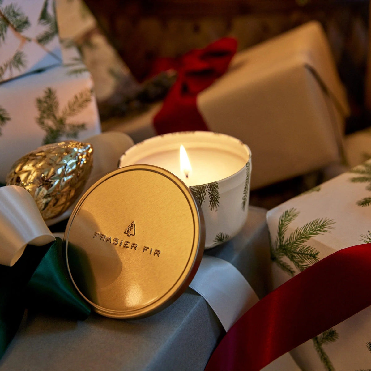 Candle with 'Frasier Fir' lid on a table with wrapped gifts and ribbons.