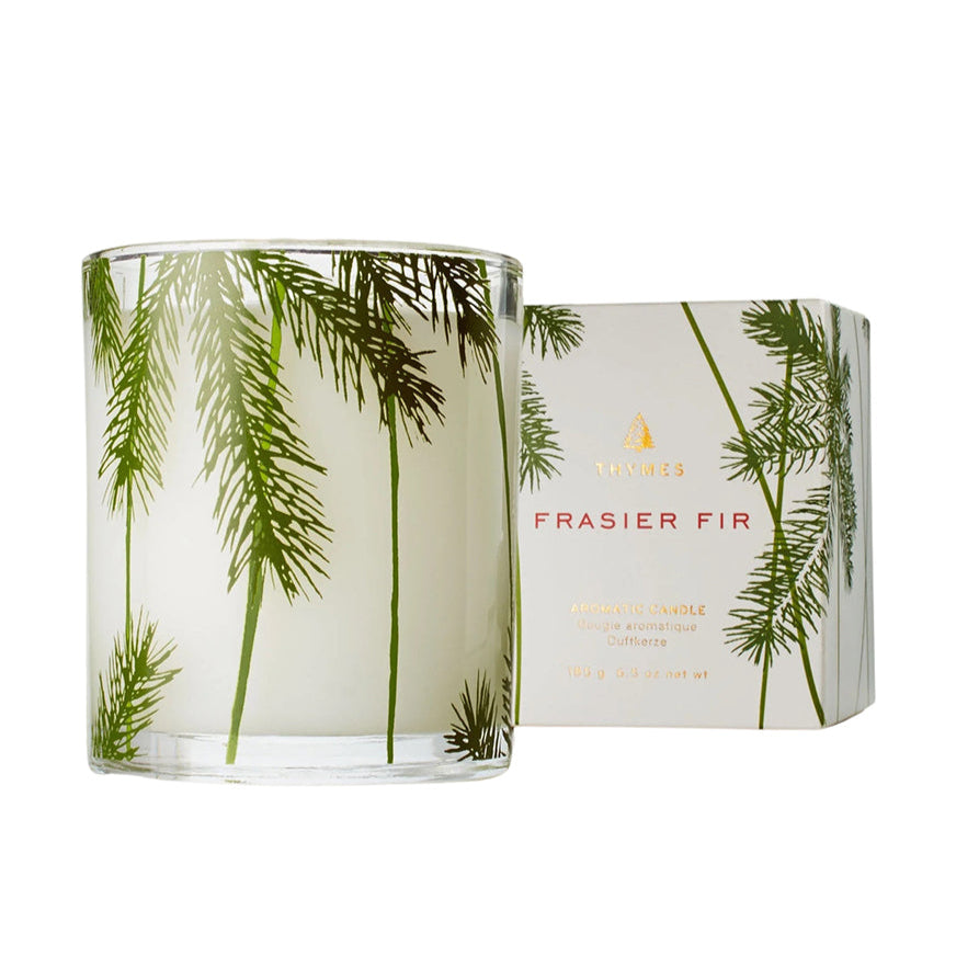 A clear candle with a design of green pine needles surrounding it, labeled 'Frasier Fir' with the Thymes brand visible on the packaging.