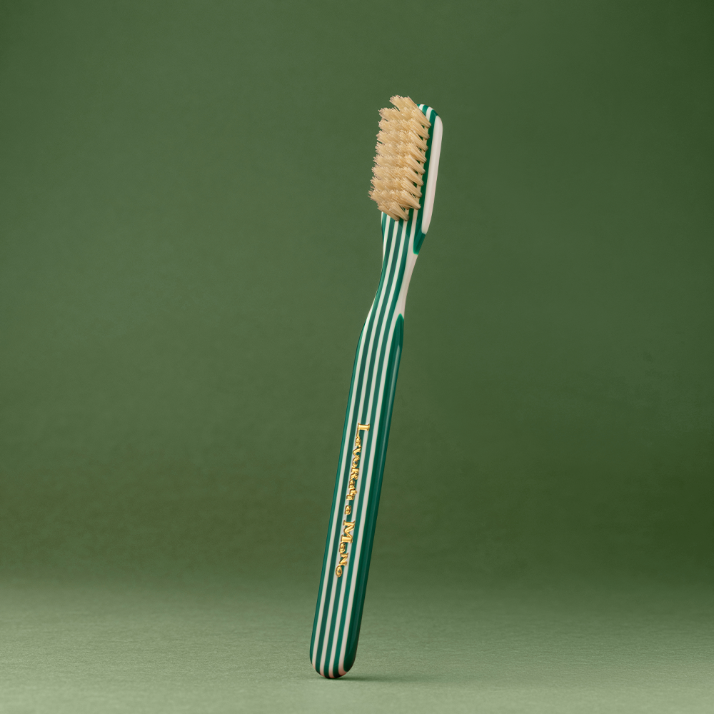 Green Capri Collection Toothbrush with a green and white striped handle and beige bristles, standing upright against a solid green background.