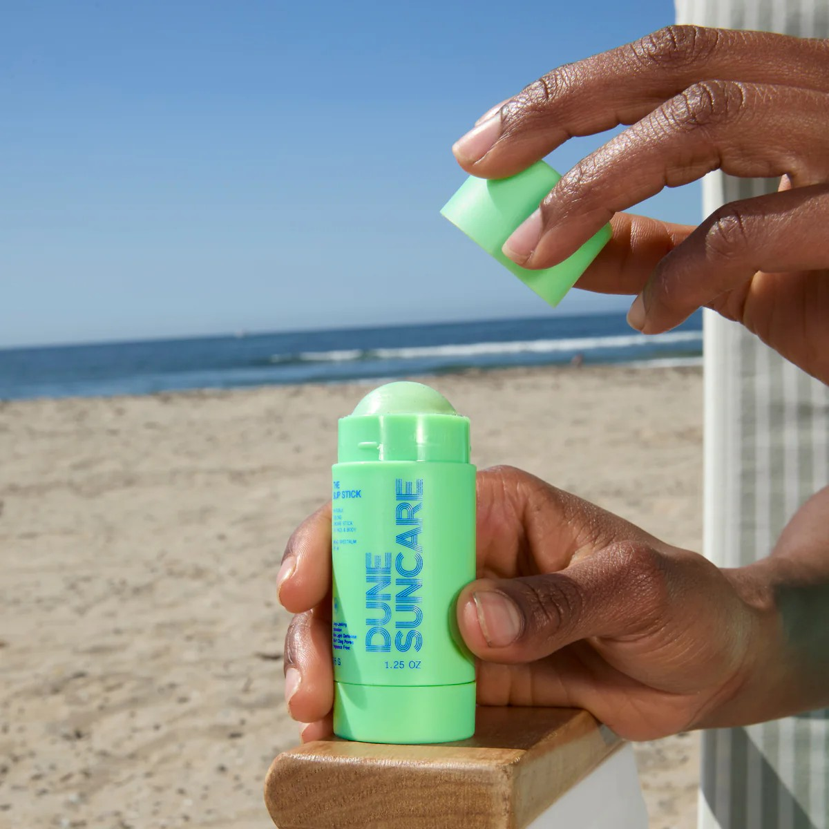 Person holding a green sunscreen stick with beach in the background