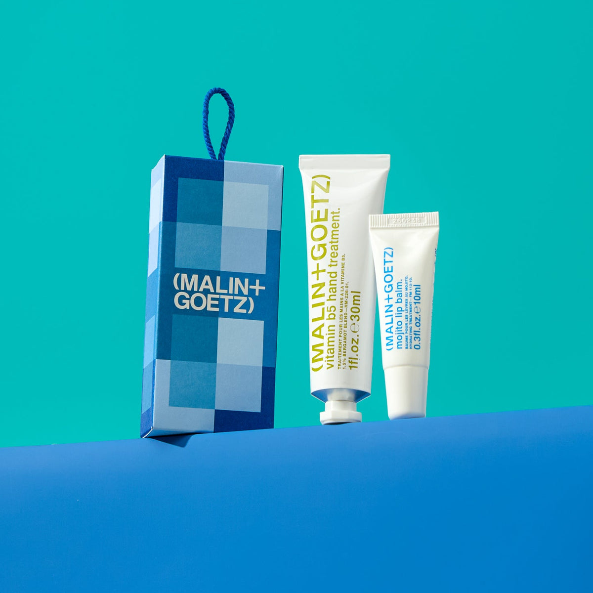 A Malin+Goetz skincare gift set featuring a blue and white checkered gift bag and two white tubes of products, displayed on a blue surface against a teal background.