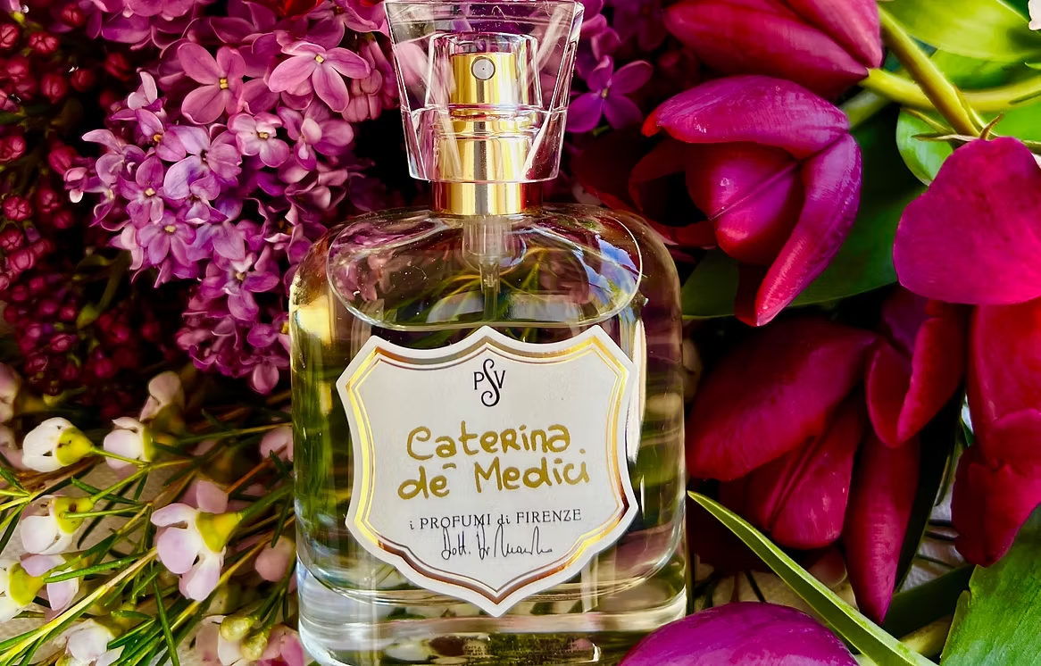 A clear glass perfume bottle labeled 'Caterina de Medici' with a gold spray top, surrounded by vibrant pink and purple flowers.