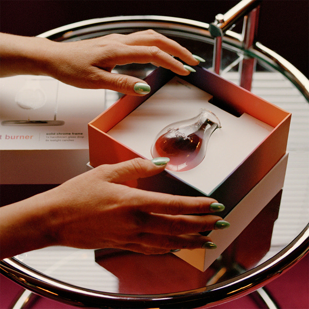 Person opening a box containing a small glass vessel on a reflective surface.