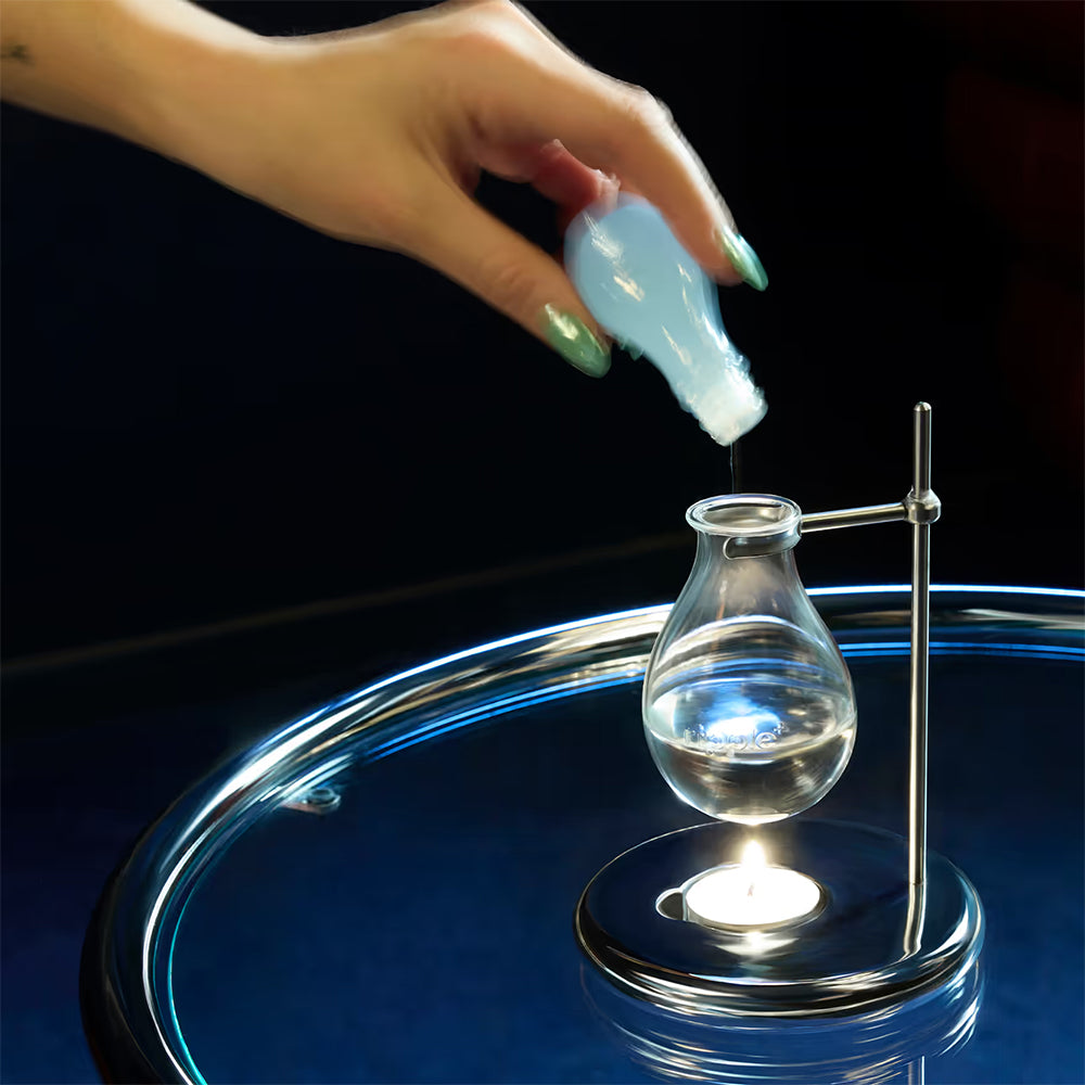 Hand pouring a liquid into a small glass container on a reflective surface with a dark background