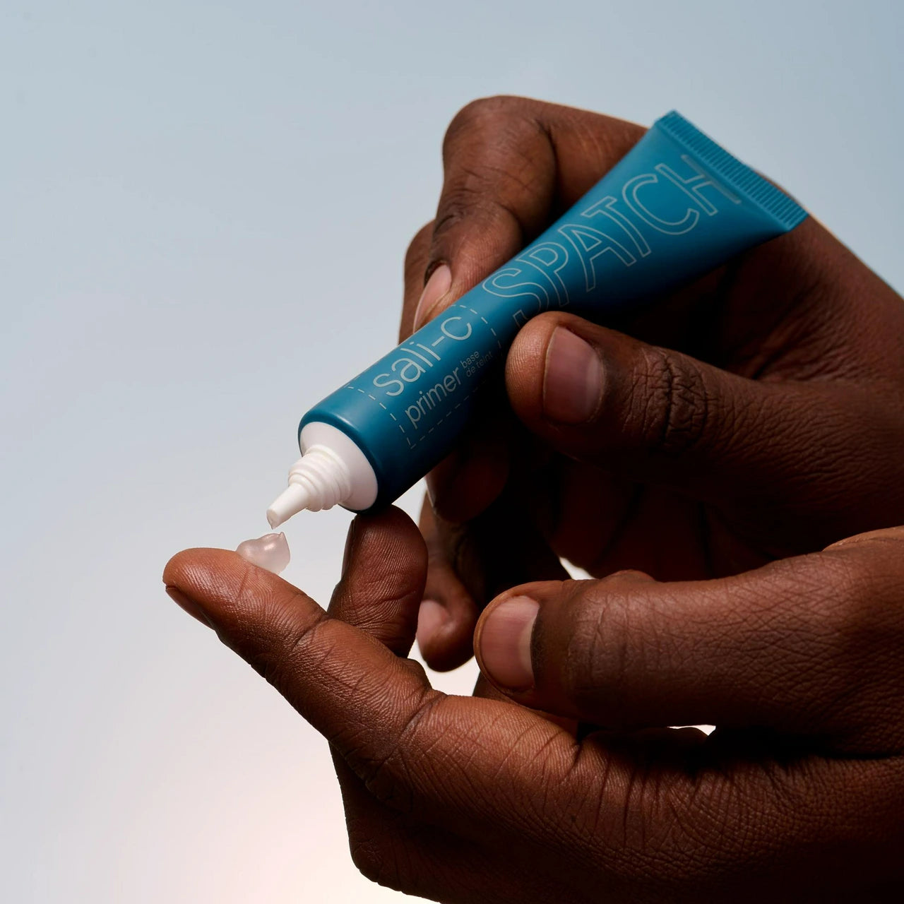 A close-up of a person's hands squeezing a teal tube of Spatch primer onto their finger, with a small clear gel-like droplet falling, against a soft light gray background.