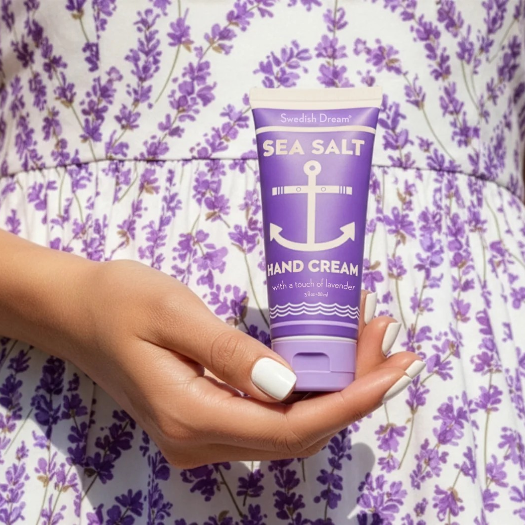 A woman's hand holding a tube of Lavender Sea Salt Hand Cream.  You can see the lavender print dress the woman is wearing as well.