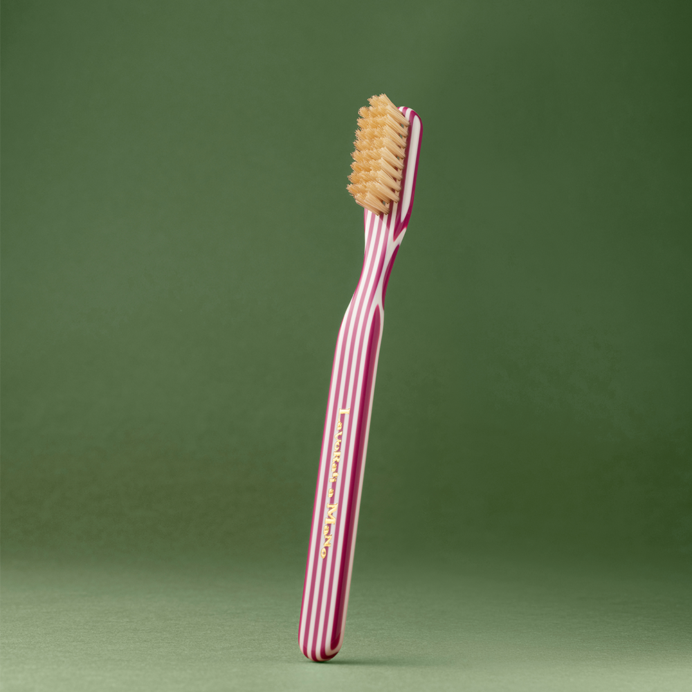Magenta Capri Collection Toothbrush with a pink and white striped handle and beige bristles, standing upright against a solid green background.
